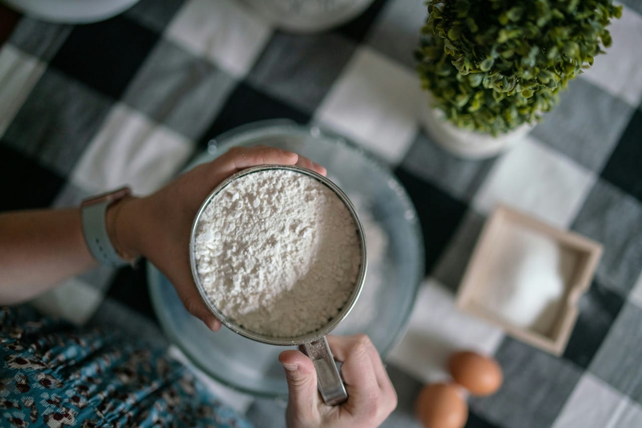 Hands holding a metal measuring cup filled with flour above a mixing bowl